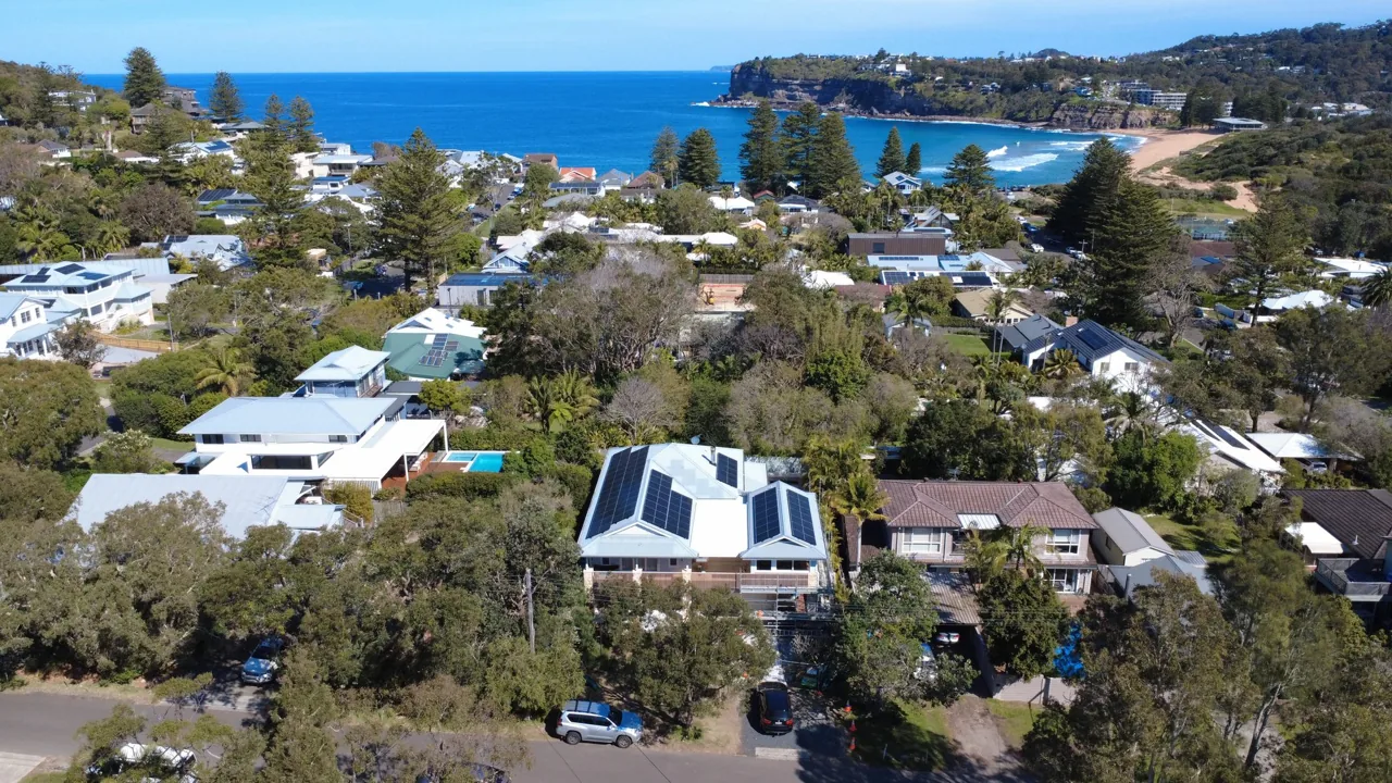 Aerial view of coastal homes with rooftop solar panels and a bright blue ocean and headland beyond