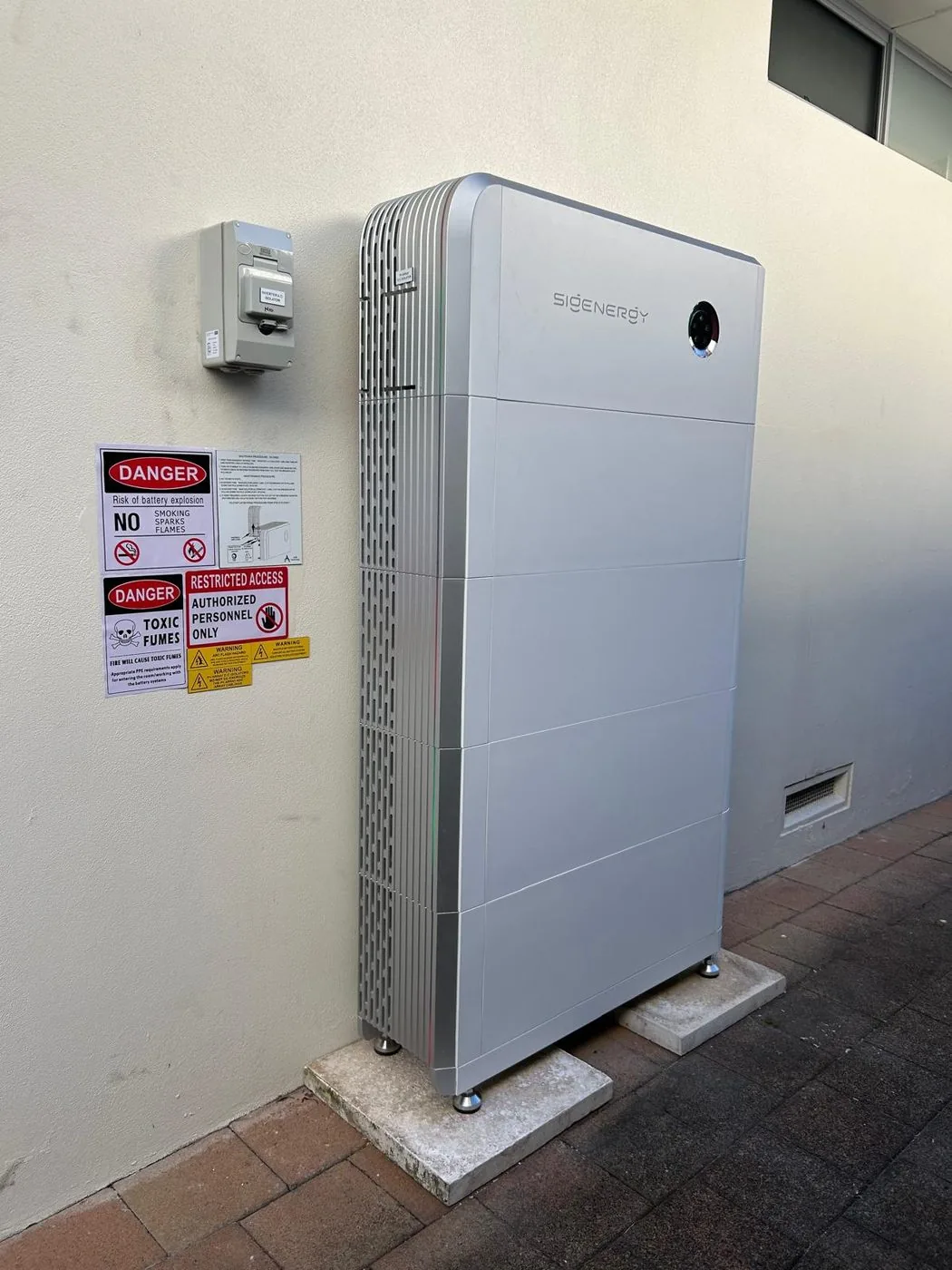 Silver Sigenergy battery stack on a paved side path beside a rendered wall